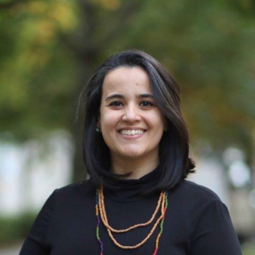 Fotografia em retrato de Natália Martins de Oliveira Gonçalves, Gerente da ELACult. Ela aparece sorrindo, de frente para a câmera, usando blusa preta de gola alta e colares longos de contas. Tem cabelos escuros, lisos, na altura dos ombros. Ao fundo, há uma paisagem externa desfocada, com tons de verde, indicando um ambiente ao ar livre.